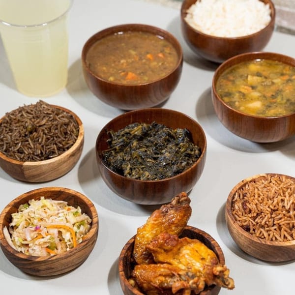 A spread of Haitian dishes including rice, soups, coleslaw, and chicken wings in wooden bowls.