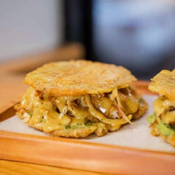 Two Blueline Fry plantain sandwiches with creamy sauce and fillings on a wooden tray.